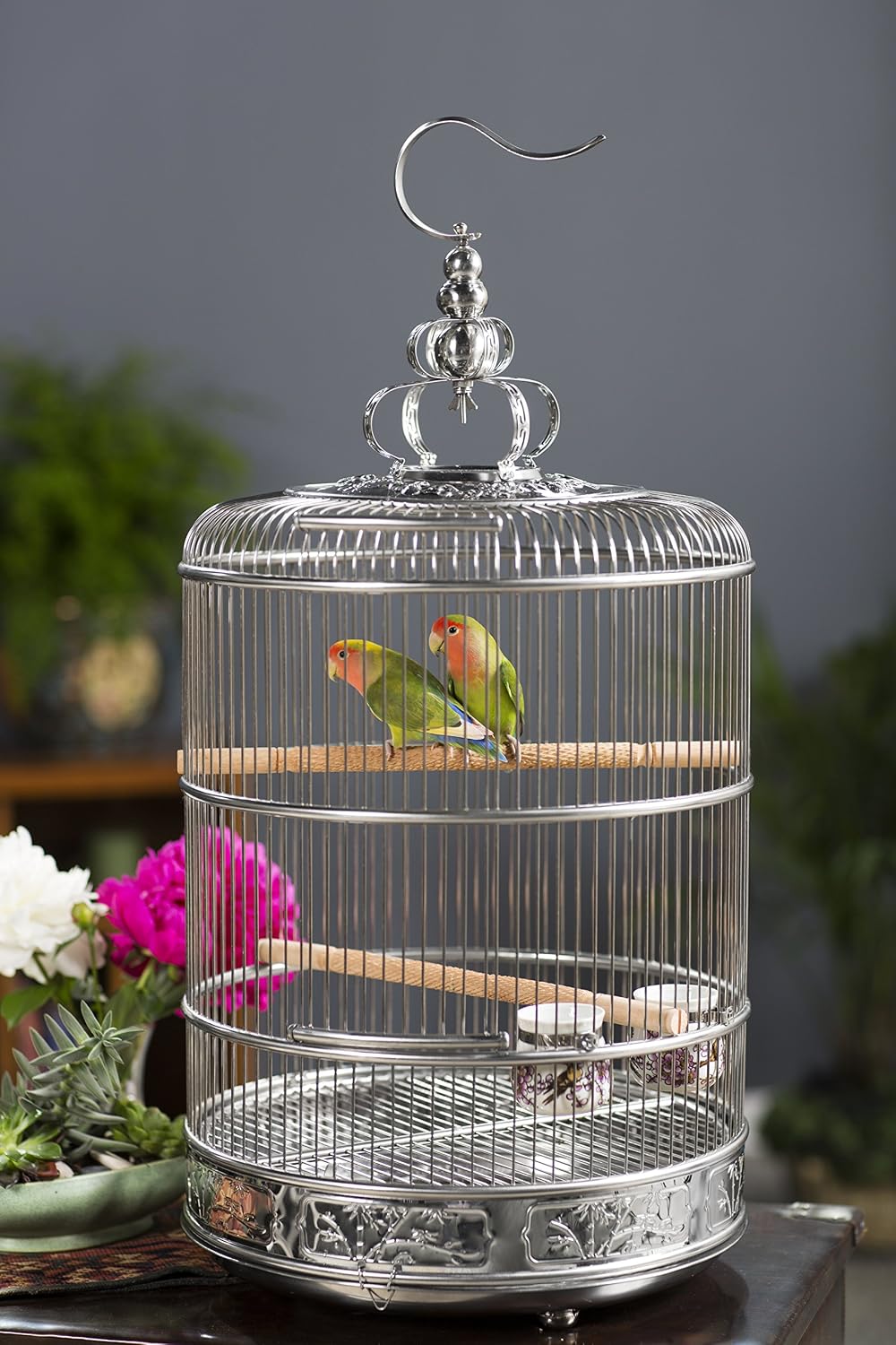 A decorative silver metal birdcage with two small green lovebirds perched inside, set against a blurred gray background with flowers and greenery.