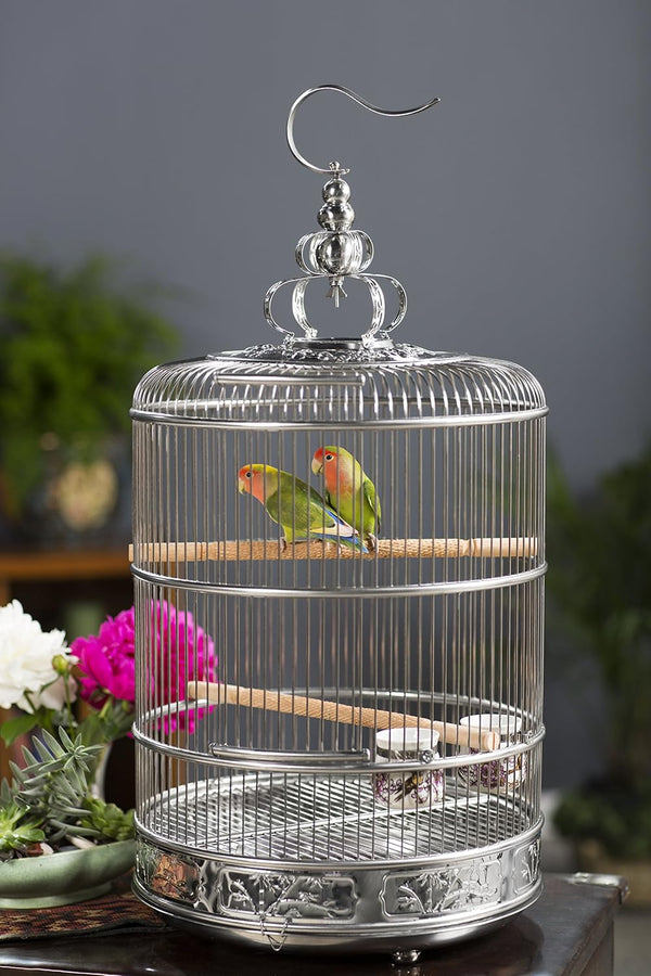 A decorative silver metal birdcage with two small green lovebirds perched inside, set against a blurred gray background with flowers and greenery.